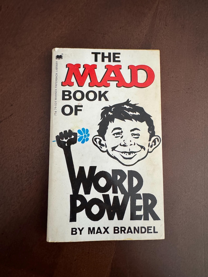 Pode incluir: Um livro de bolso vintage intitulado "The Mad Book of Word Power" de Max Brandel. A capa apresenta uma ilustra&ccedil;&atilde;o de desenho animado de um rosto sorridente e um punho erguido segurando uma flor. O t&iacute;tulo &eacute; em vermelho e preto.