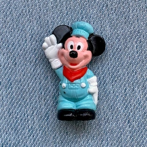 May include: A small plastic figurine of Mickey Mouse wearing a blue and white uniform with a red bandana. He is waving with his right hand and has a black hat.