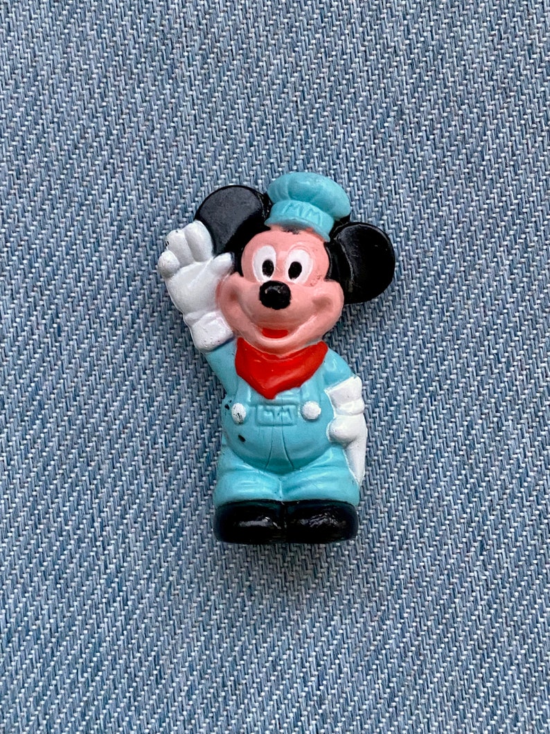 May include: A small plastic figurine of Mickey Mouse wearing a blue and white uniform with a red bandana. He is waving with his right hand and has a black hat.