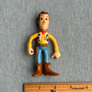 May include: A Woody action figure, featuring a yellow shirt, blue pants, and brown boots. The figure has a sheriff's badge and a red neckerchief. A wooden ruler with inch measurements is in the foreground.