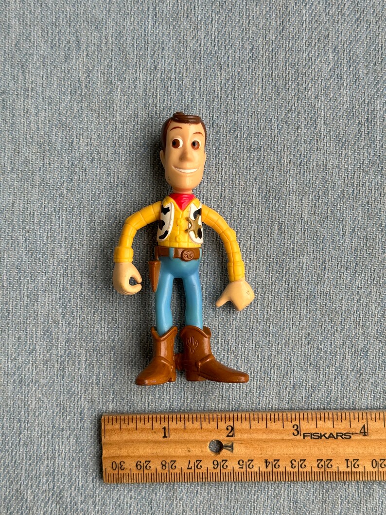 May include: A Woody action figure, featuring a yellow shirt, blue pants, and brown boots. The figure has a sheriff's badge and a red neckerchief. A wooden ruler with inch measurements is in the foreground.