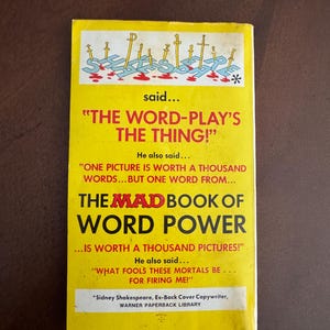 Pode incluir: Uma capa de livro de bolso vintage com um fundo amarelo brilhante. A capa apresenta o t&iacute;tulo "THE MAD BOOK OF WORD POWER" em letras grandes e em negrito. A capa tamb&eacute;m inclui o texto que diz "THE WORD-PLAY'S THE THING!" e "ONE PICTURE IS WORTH A THOUSAND WORDS..."