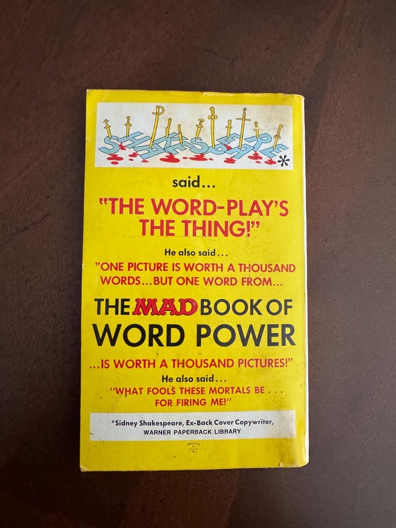 Pode incluir: Uma capa de livro de bolso vintage com um fundo amarelo brilhante. A capa apresenta o t&iacute;tulo "THE MAD BOOK OF WORD POWER" em letras grandes e em negrito. A capa tamb&eacute;m inclui o texto que diz "THE WORD-PLAY'S THE THING!" e "ONE PICTURE IS WORTH A THOUSAND WORDS..."