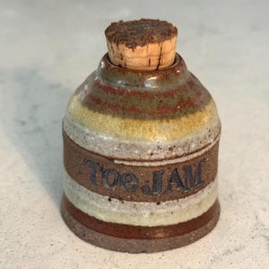May include: A handmade ceramic jar with a cork stopper. The jar is cylindrical with horizontal bands of brown, yellow, and white. The words "Toe Jam" are inscribed on the side. The jar is likely used for storage.