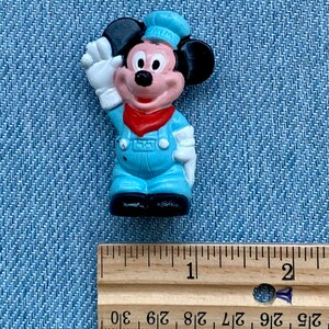 May include: A small plastic figurine of Mickey Mouse wearing a blue and white conductor's uniform. He is waving with his right hand and has a red bandana around his neck. The figurine is approximately 2.5 cm tall.
