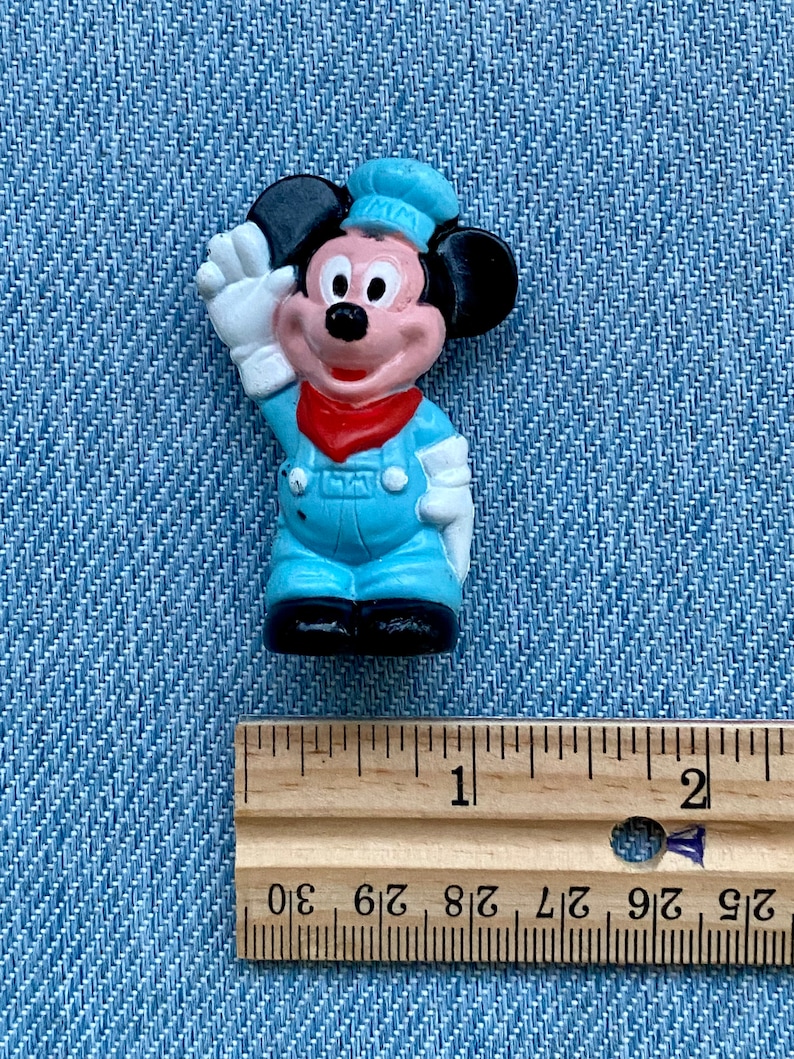 May include: A small plastic figurine of Mickey Mouse wearing a blue and white conductor's uniform. He is waving with his right hand and has a red bandana around his neck. The figurine is approximately 2.5 cm tall.