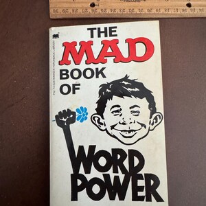 Pode incluir: Um livro de bolso vintage intitulado "The Mad Book of Word Power" de Max Brandel. A capa apresenta uma ilustra&ccedil;&atilde;o de desenho animado de um menino sorrindo e um punho erguido segurando uma flor. Uma r&eacute;gua de madeira &eacute; colocada acima do livro.