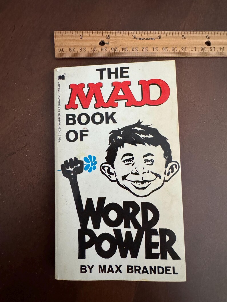 Pode incluir: Um livro de bolso vintage intitulado "The Mad Book of Word Power" de Max Brandel. A capa apresenta uma ilustra&ccedil;&atilde;o de desenho animado de um menino sorrindo e um punho erguido segurando uma flor. Uma r&eacute;gua de madeira &eacute; colocada acima do livro.