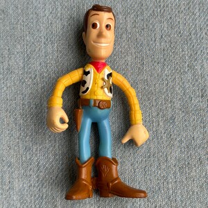 May include: A Woody action figure from the Toy Story franchise. The figure is wearing a yellow shirt, blue pants, and brown cowboy boots. It has a red neckerchief and a sheriff's badge.