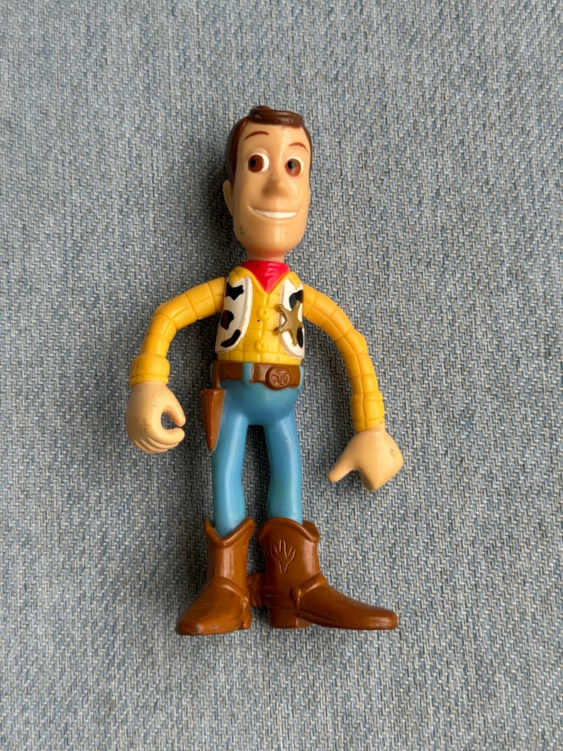 May include: A Woody action figure from the Toy Story franchise. The figure is wearing a yellow shirt, blue pants, and brown cowboy boots. It has a red neckerchief and a sheriff's badge.