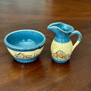 May include: A small, blue and yellow ceramic bowl and creamer set. The bowl and creamer feature a hand-painted design of a cabin and trees. The creamer has a handle and spout. The set is likely vintage or handmade.