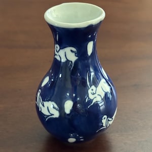 May include: A small, blue and white ceramic vase with a rounded body and a narrow neck. The vase is decorated with a white design of rabbits and dots.
