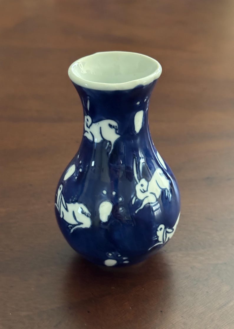 May include: A small, blue and white ceramic vase with a rounded body and a narrow neck. The vase is decorated with a white design of rabbits and dots.
