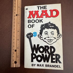 Pode incluir: Um livro de bolso vintage intitulado "The Mad Book of Word Power" de Max Brandel. A capa apresenta uma ilustra&ccedil;&atilde;o de desenho animado de um rosto sorridente e um punho segurando uma flor. Uma r&eacute;gua de madeira est&aacute; ao lado do livro.