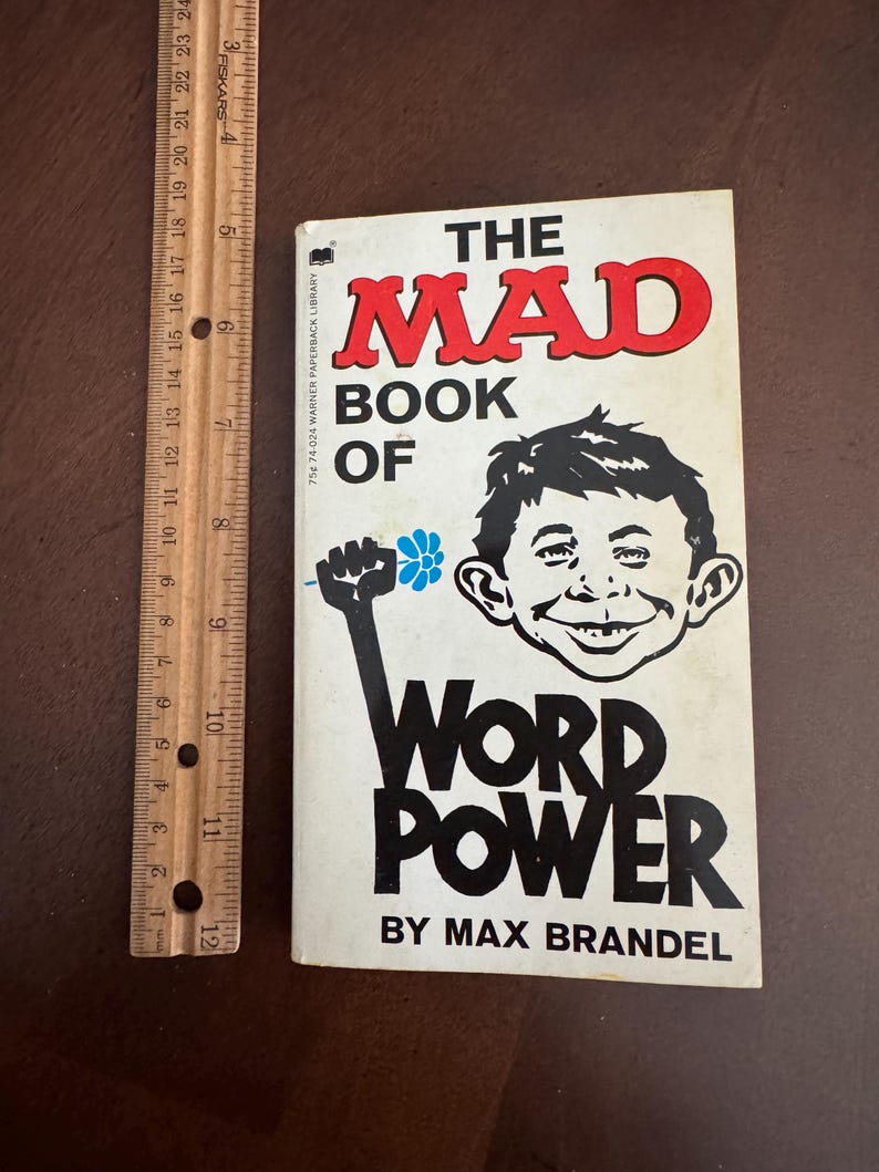 Pode incluir: Um livro de bolso vintage intitulado "The Mad Book of Word Power" de Max Brandel. A capa apresenta uma ilustra&ccedil;&atilde;o de desenho animado de um rosto sorridente e um punho segurando uma flor. Uma r&eacute;gua de madeira est&aacute; ao lado do livro.