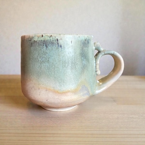 May include: A handmade ceramic mug with a rounded base and a curved handle. The mug features a gradient of colors, transitioning from a light pink at the base to a pale green at the top, with hints of brown and yellow. The mug is sitting on a wooden surface.
