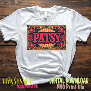 May include: White t-shirt with a colorful graphic that says "PATSY" in red letters. The graphic is framed by a decorative border with flowers and a sunburst design. The shirt is knotted at the bottom.