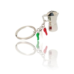 Puede incluir: Un llavero de color plateado con un dije en miniatura de cafetera. El dije está acompañado de tres dijes de pimientos en los colores de la bandera italiana: verde, blanco y rojo. El llavero está sobre un fondo blanco.