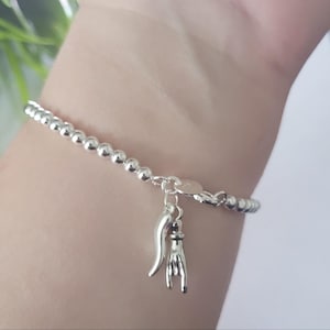 May include: A silver beaded bracelet featuring two dangling charms: a curved horn-shaped pendant and a hand gesture charm depicting the sign of the horns. The bracelet is constructed from small, uniform, polished silver spheres linked together.