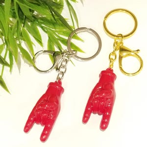 May include: Two keychains with red hand pendants in the 'rock on' gesture. One keychain has a silver ring and chain, the other has a gold ring and clasp. Green foliage is in the background.