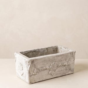 May include: A rectangular planter with a weathered, off-white finish. The exterior features raised, ornate floral relief detailing along the front and sides, with a rough, textured surface throughout the entire structure of the container.