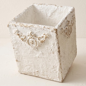 May include: A square-shaped container with a rough, textured white finish and distressed edges. The exterior features raised, ornate gold-toned scrollwork detailing on the sides, providing a decorative accent to the otherwise plain, rugged surface.