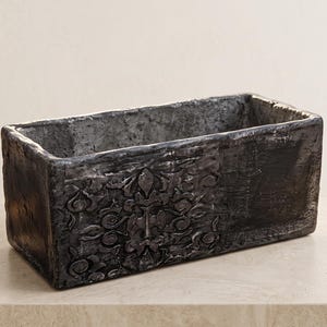 May include: A rectangular planter with a dark, weathered charcoal finish. The exterior features a raised, symmetrical floral relief pattern on the front face, while the interior and sides display a rough, textured surface with uneven edges.