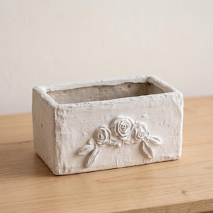 May include: A rectangular white planter with a textured, matte finish. The front side features a raised, sculpted relief of three roses with leaves arranged in a horizontal cluster. The container has thick walls and an open top for holding items.