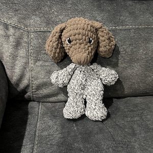 May include: A brown and white crocheted stuffed dog toy sitting on a gray couch.