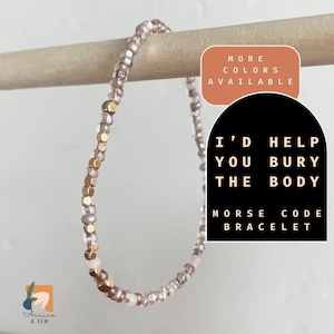 May include: A pink and gold beaded bracelet with the message "I'd help you bury the body" in Morse code. The bracelet is displayed on a wooden surface with a sign that says "More colors available".
