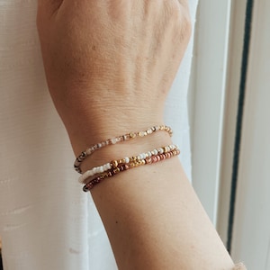 May include: Three beaded bracelets stacked on a wrist. The top bracelet is made of light pink and gold beads. The middle bracelet is made of white and gold beads. The bottom bracelet is made of brown and gold beads.