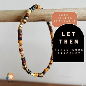 May include: A multi-colored beaded bracelet with gold accents. The bracelet is made of small, round beads in shades of brown, black, cream, and gold. The bracelet is displayed on a wooden surface. The text "LET THEM" and "MORSE CODE BRACELET" is displayed on a black background.