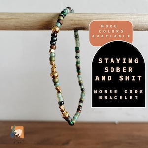 May include: A multi-colored beaded bracelet with gold accents. The bracelet is on a wooden surface and has a sign that reads "Staying Sober and Shit Morse Code Bracelet".