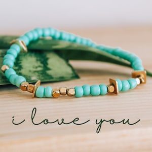 May include: A turquoise beaded bracelet with gold accents. The bracelet is on a green leaf background. The text "i love you" is written below the bracelet.