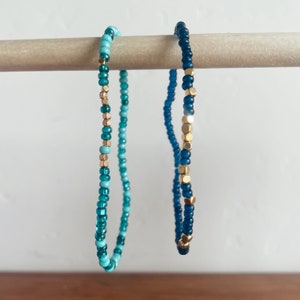 May include: Two beaded bracelets with a gradient of blue and turquoise beads. One bracelet has gold beads interspersed throughout.