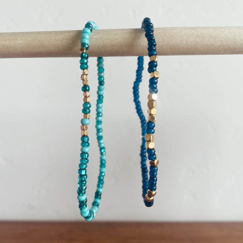 May include: Two beaded bracelets with a gradient of blue and turquoise beads. One bracelet has gold beads interspersed throughout.