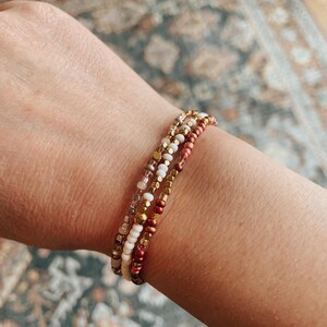 May include: A set of three beaded bracelets in shades of brown, white, and gold. The bracelets are made with small, round beads and are stacked on a person's wrist.
