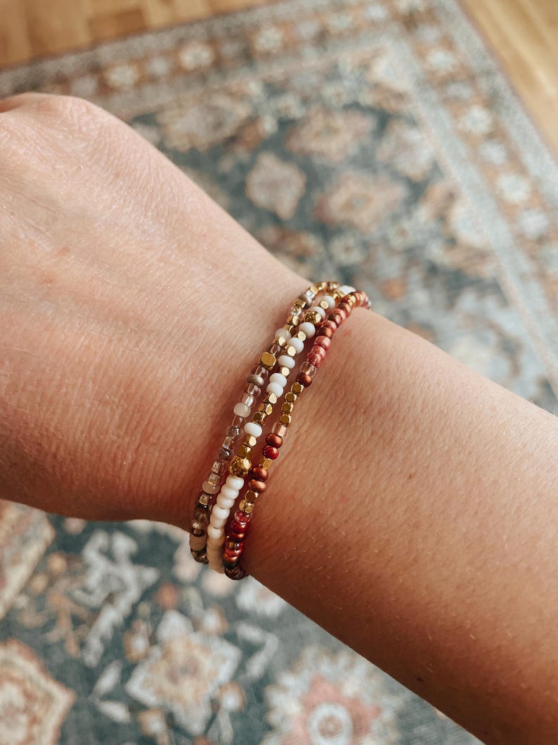 May include: A set of three beaded bracelets in shades of brown, white, and gold. The bracelets are made with small, round beads and are stacked on a person's wrist.
