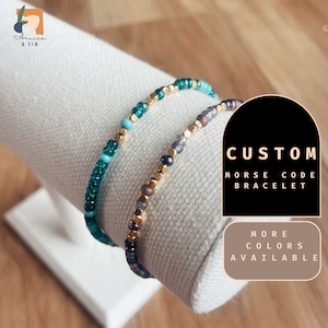May include: Two custom Morse code bracelets, one with teal and turquoise beads and gold accents, and the other with gray and black beads and gold accents.  The bracelets are on a white display stand.  "CUSTOM MORSE CODE BRACELET" and "MORE COLORS AVAILABLE" are written in black text on a white background.