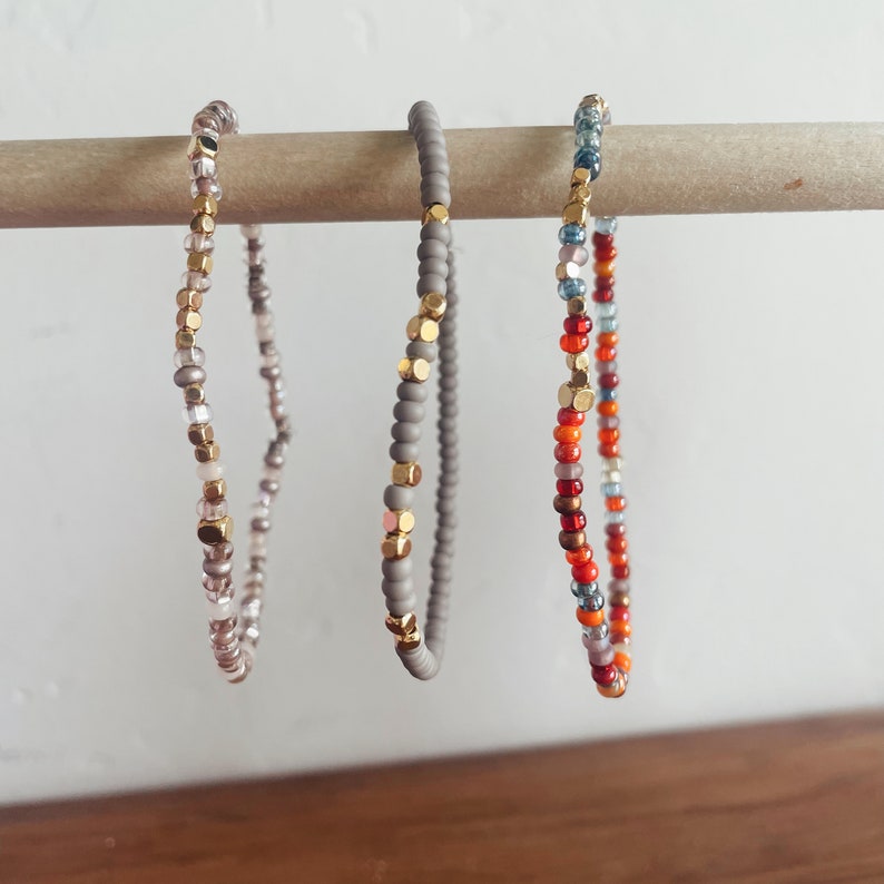 May include: Three beaded bracelets with gold accents. The bracelets are displayed on a wooden dowel. The bracelets are made with gray, white, and pink beads, gray beads, and red, orange, and blue beads.