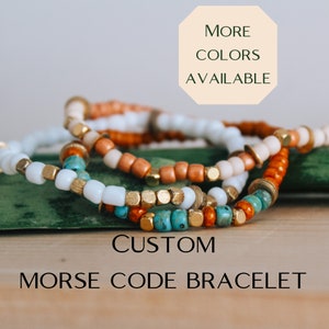 May include: A set of custom Morse code bracelets with white, orange, turquoise, and gold beads. The bracelets are stacked on top of each other and are on a wooden surface. The text "CUSTOM MORSE CODE BRACELET" is below the bracelets. The text "MORE COLORS AVAILABLE" is in a peach-colored hexagon above the bracelets.