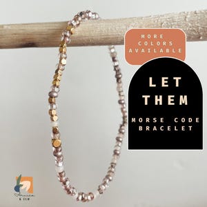 May include: A delicate gold and pink beaded bracelet with the words "LET THEM" in Morse code. The bracelet is on a wooden surface and the text "MORE COLORS AVAILABLE" is in a circle above the bracelet.