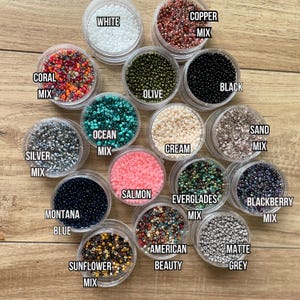 May include: Assortment of 14 small containers filled with various colors of seed beads. The containers are labeled with the bead color names, such as "Coral Mix", "Silver Mix", "Montana Blue", "Copper Mix", "White", "Black", "Olive", "Ocean Mix", "Cream", "Salmon", "Everglades Mix", "Sunflower Mix", "American Beauty", "Matte Grey", "Sand Mix", and "Blackberry Mix".