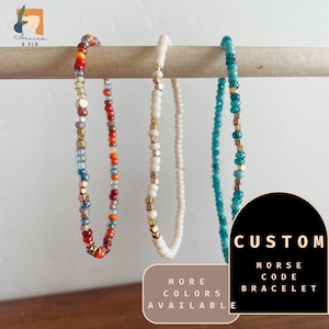 May include: Three beaded bracelets with gold accents. The bracelets are in red, white, and teal colors. The text "CUSTOM MORSE CODE BRACELET" is in a black circle with a white outline. The text "MORE COLORS AVAILABLE" is in a white circle with a black outline.