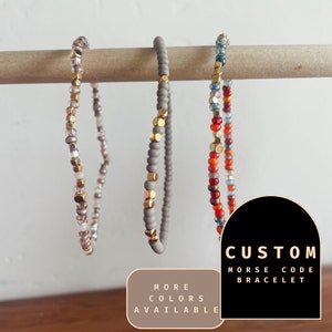May include: Three beaded bracelets in different color combinations, including gray, white, gold, red, orange, and blue. The bracelets are hanging on a wooden dowel rod. The text "CUSTOM MORSE CODE BRACELET" is displayed on a black background with a white outline. The text "MORE COLORS AVAILABLE" is displayed on a brown background with a white outline.
