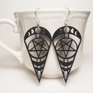 Puede incluir: Un par de pendientes negros colgantes con diseños de pentagrama, luna creciente y círculo. Los pendientes cuelgan de una taza de cerámica blanca.