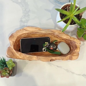 May include: A wooden, freeform bowl in a light brown hue contains a black smartphone, car keys, and a small, smooth, grey stone. The bowl's organic shape and colour create a rustic aesthetic. The items are arranged on a white surface, with potted succulents in the background.