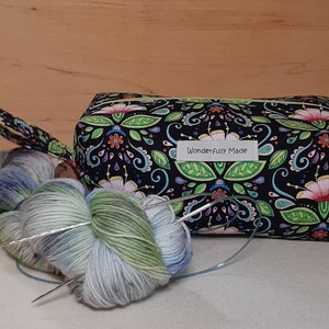 May include: A black fabric project bag with a floral pattern and a zipper closure. The bag has a green strap and a label that reads "Wonderfully Made". Two balls of yarn and knitting needles are in front of the bag.