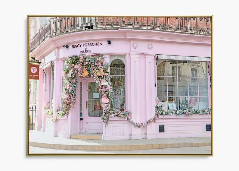 May include: A pink storefront with a sign that reads "Peggy Porschen Cakes" decorated with flowers and greenery. The storefront has a large glass window and a door with a floral wreath.