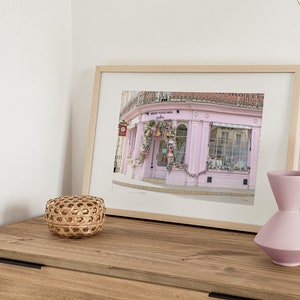 May include: A framed print of a pink storefront with a sign that reads "MERRY PUCKLECHURCH Indies". The storefront is decorated with flowers and greenery. The print is on a wooden table with a wicker basket and two pink vases.